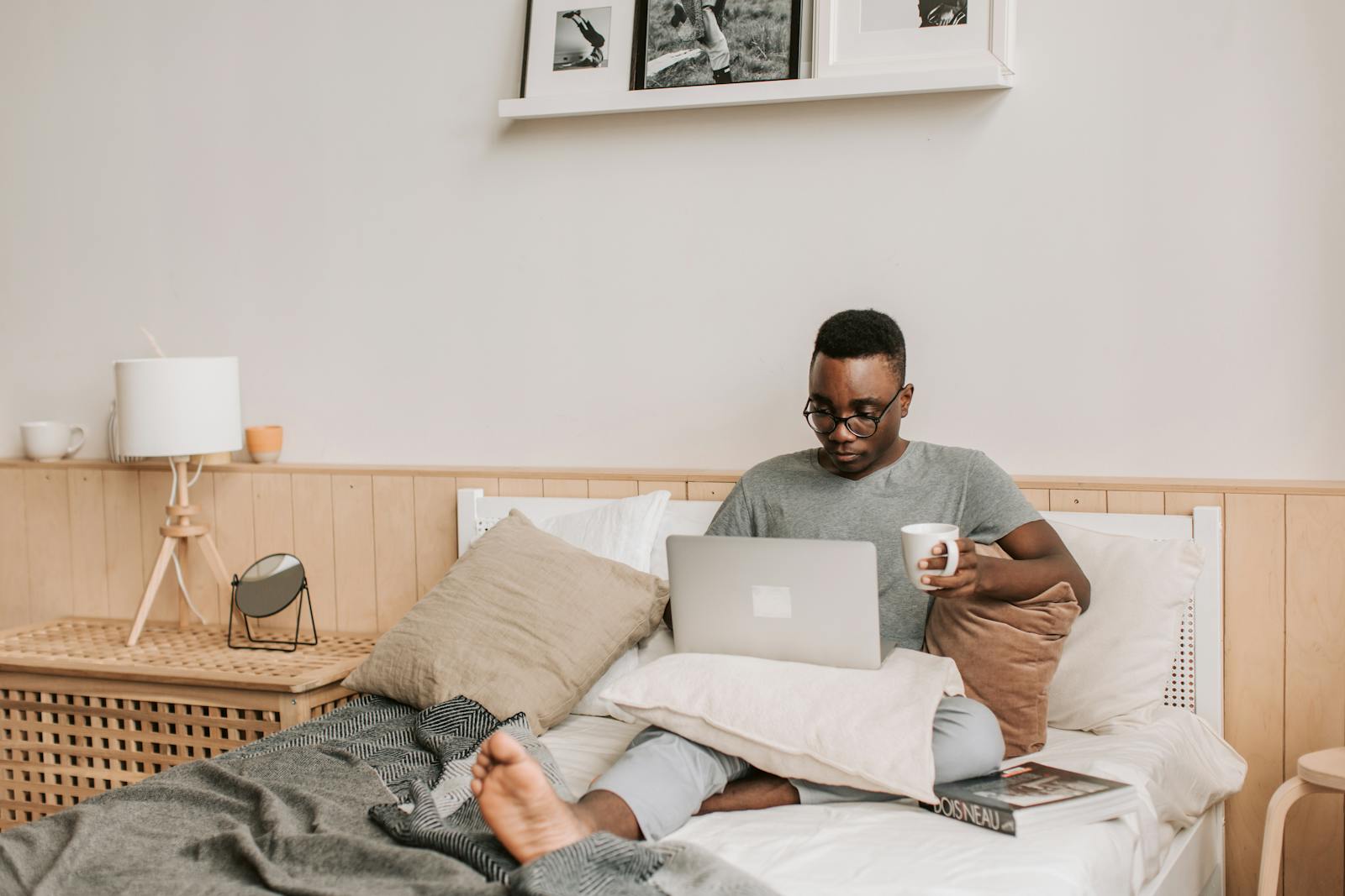 Black man sitting on a bed using a laptop for remote work with coffee in hand.